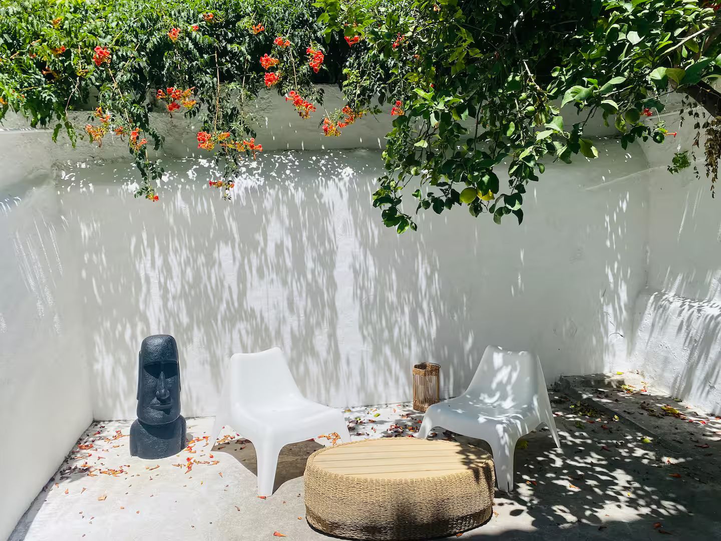 A patio area with white walls, a stone sculpture, two white plastic chairs, and a woven stool.