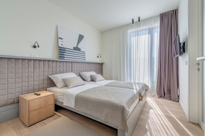 Quarto moderno com cama grande, mesa de cabeceira, TV fixada na parede e janela com cortinas.