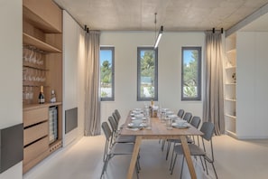A modern dining room with a long wooden table set for six, surrounded by grey chairs. There are three windows with curtains, a built-in shelf with wine glasses and a bottle, and a small built-in fridge. - Luxury 4-Bedroom Villa in Primošten with Private Pool & Panoramic Sea Views (Primošten)