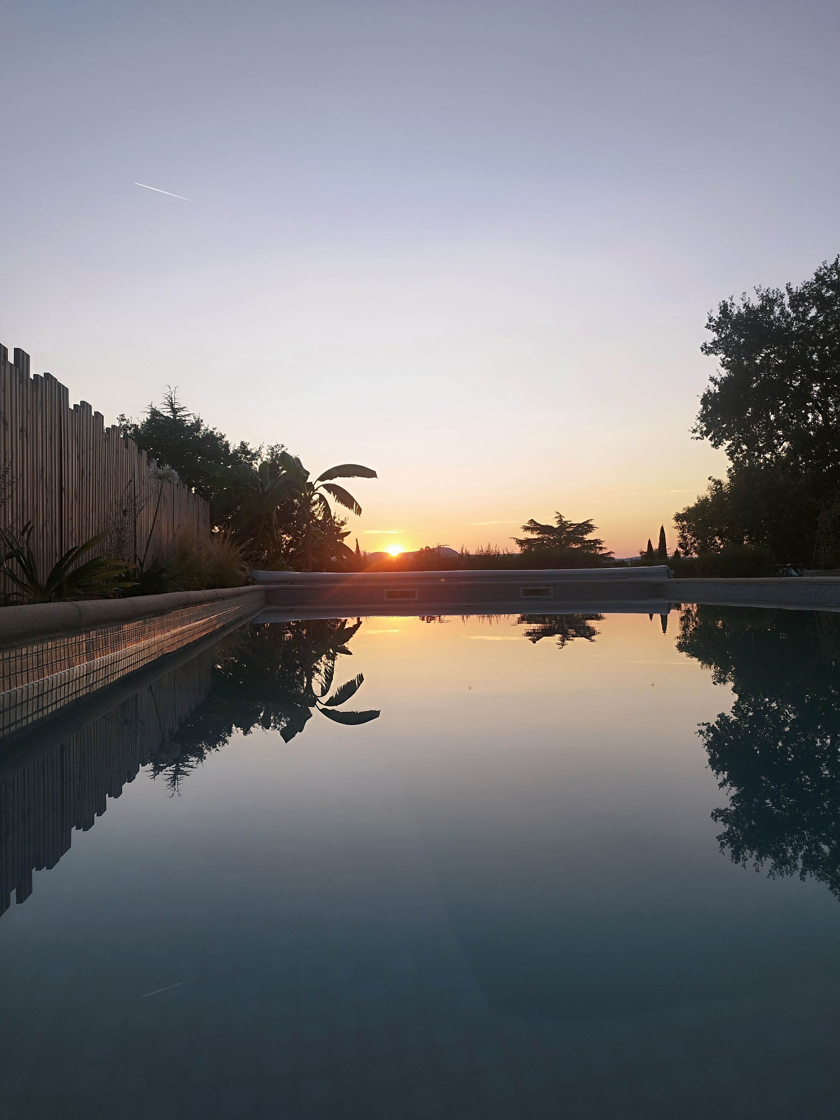 A tranquil pool reflecting the sunset, surrounded by a wooden fence and trees.