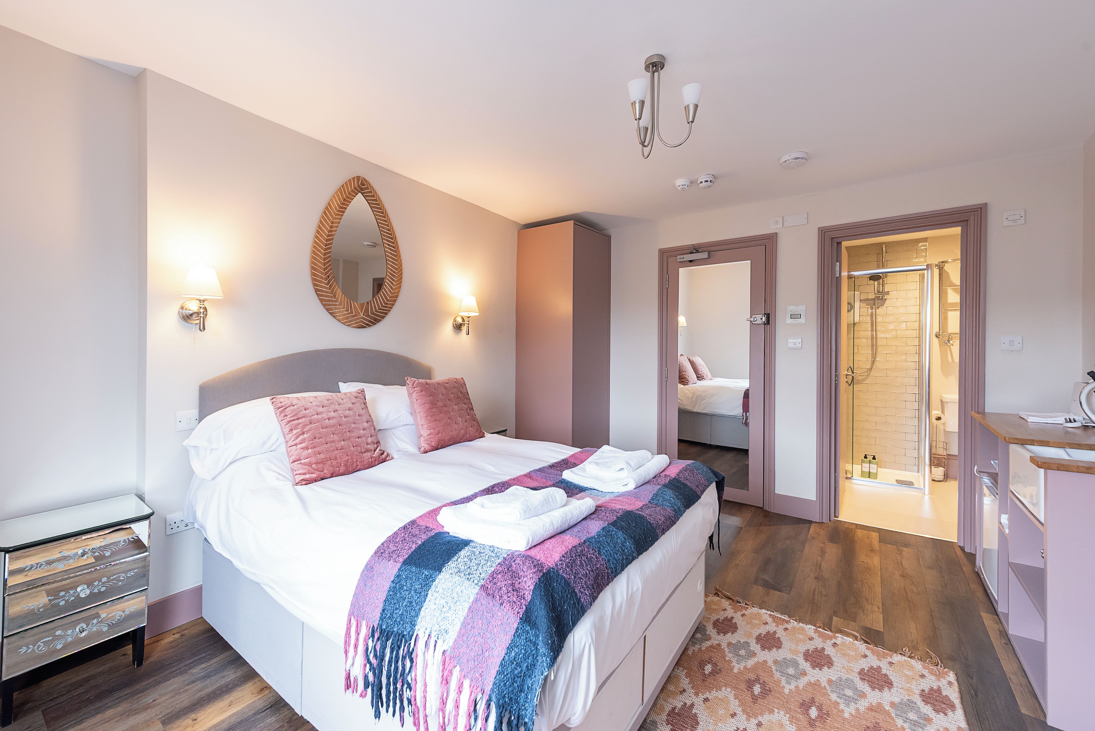 A hotel room with a bed, bedside lamps, a mirror, a bathroom with a shower, and a small desk with a chair.