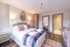 A hotel room with a bed, bedside lamps, a mirror, a bathroom with a shower, and a small desk with a chair. - Wonderous Kensington Studio (London)