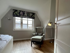 A bedroom with a checkered armchair, a window with a floral valance, a floor lamp, and a door. - Holiday Home in Nieblum With Swimming Pool (Nieblum)