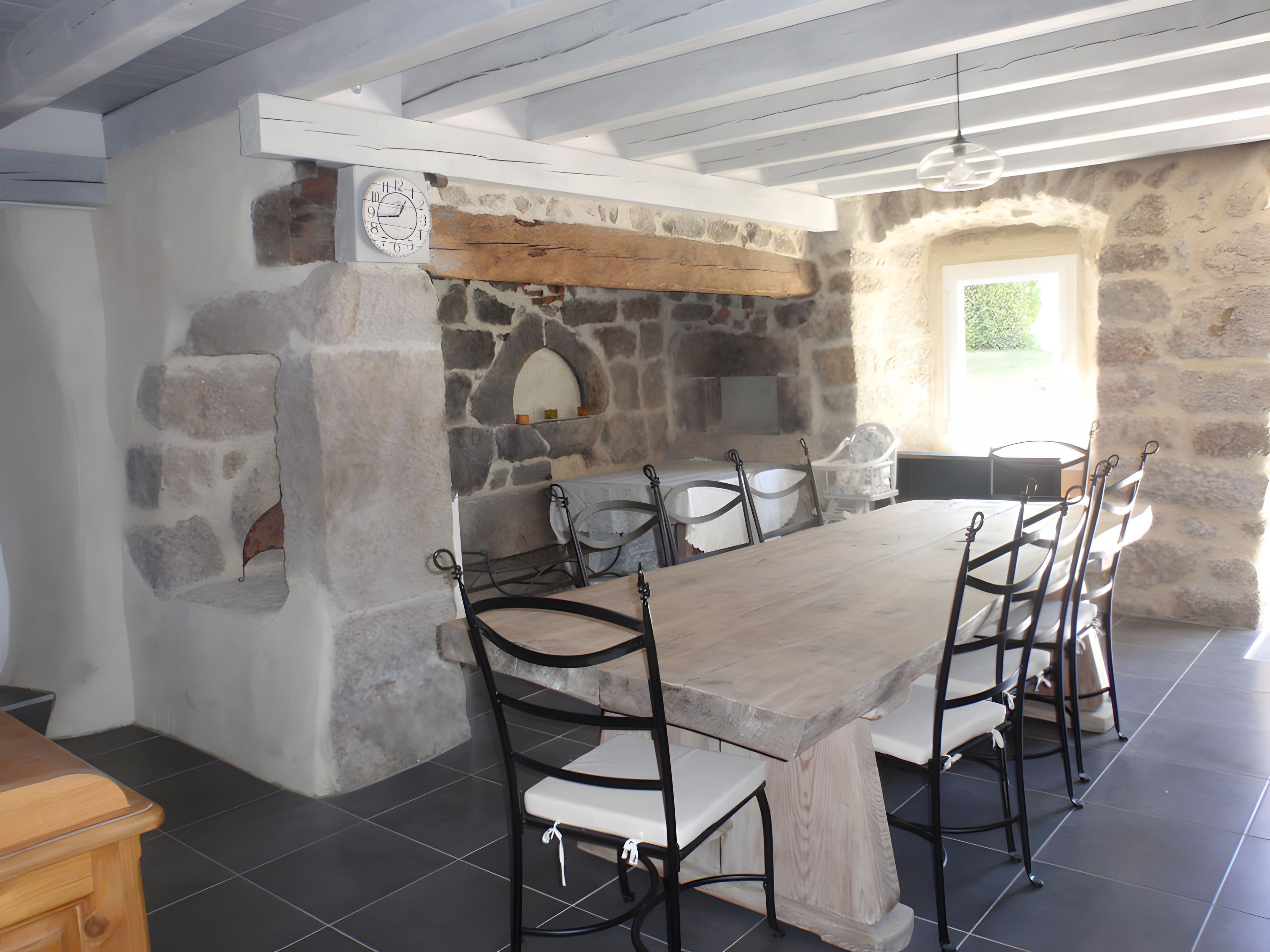 A stone-walled room with a long wooden table and metal chairs, a clock, and a window with a view.