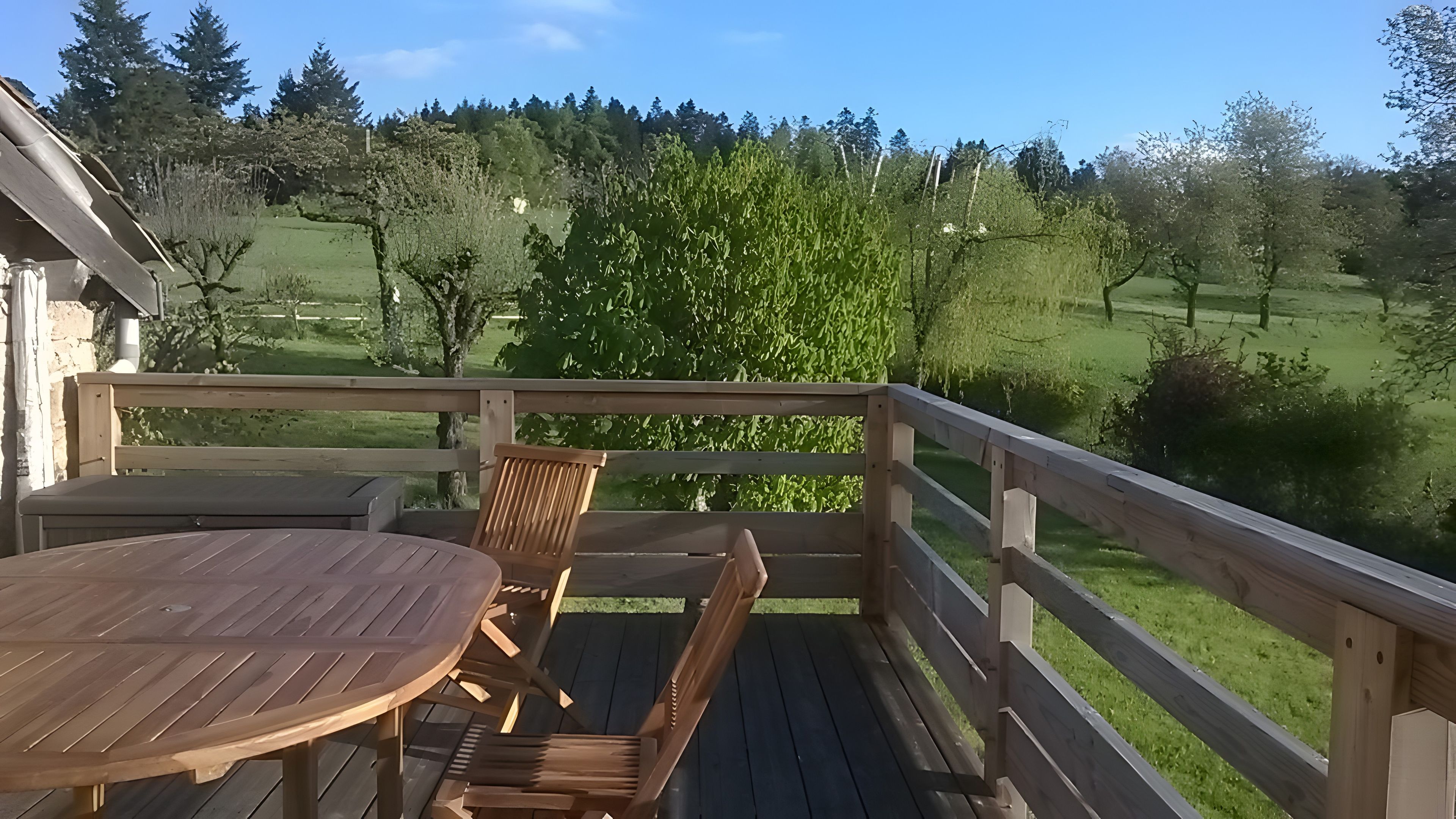 Una terraza de madera con una mesa y sillas, con vistas a un paisaje verde con árboles y cielo despejado.
