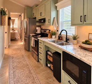 A kitchen with light green cabinets, a marble countertop, stainless steel appliances, and a window with blinds. - The Blue Door: a Be Free Tiny Home (Jacksonville)