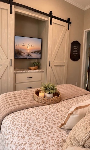 Une chambre à coucher avec un lit, un téléviseur posé sur une étagère, un panneau fixé au mur et une porte.