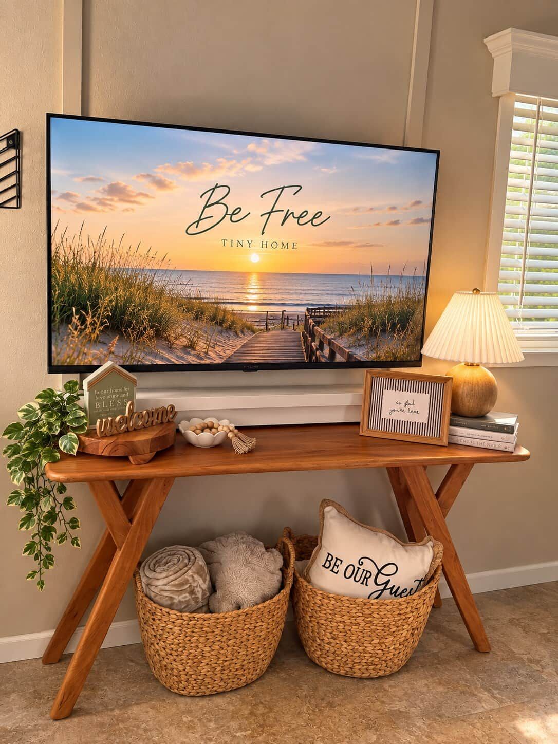 A television displaying a beach scene with the text 'Be Free Tiny Home' and a wooden console table with decorative items.