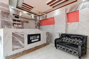 A hotel reception area with a marble reception desk, a check-out sign, a company-serviced sign, a sofa, and a ceiling fan.