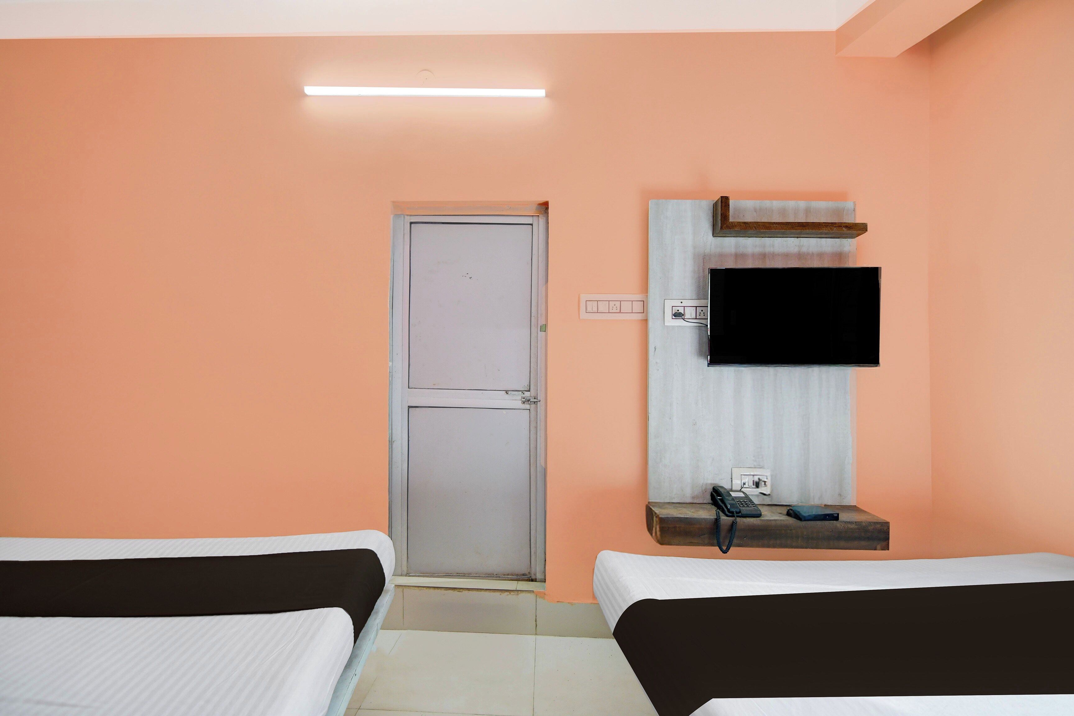 Ein Hotelzimmer mit zwei Betten, einem an der Wand befestigten Fernseher, einem Nachttisch mit Telefon und Steckdose sowie einer Tür mit Griff.