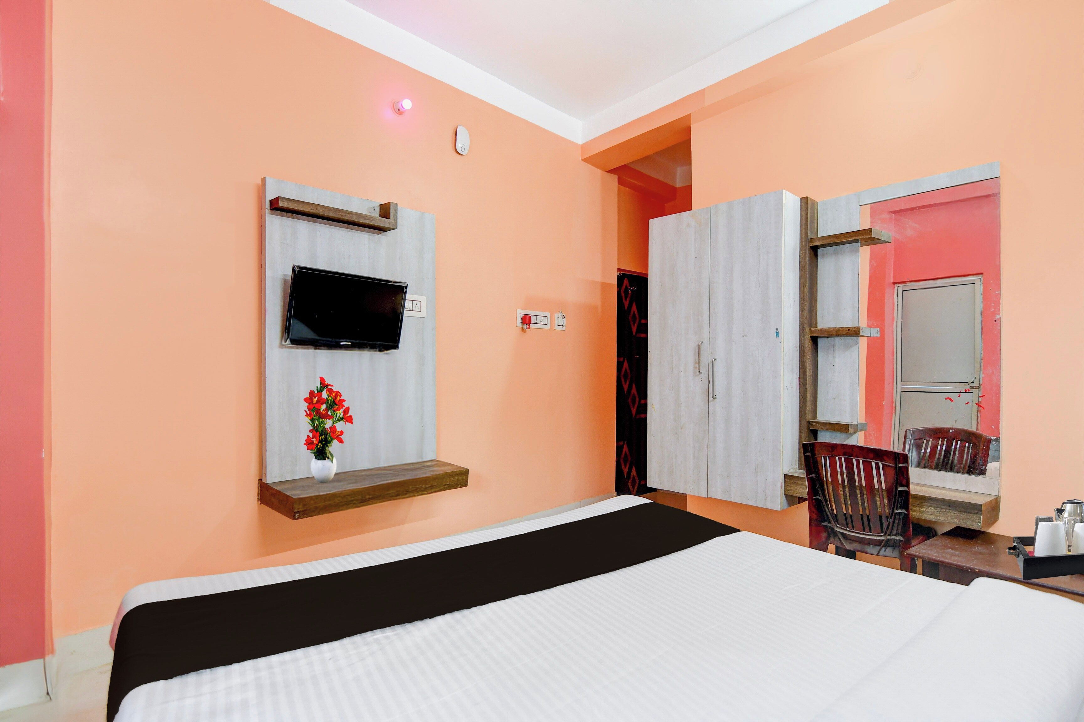 Une chambre d’hôtel avec un lit, un téléviseur fixé au mur, une petite étagère avec un vase de fleurs, une armoire et un miroir.