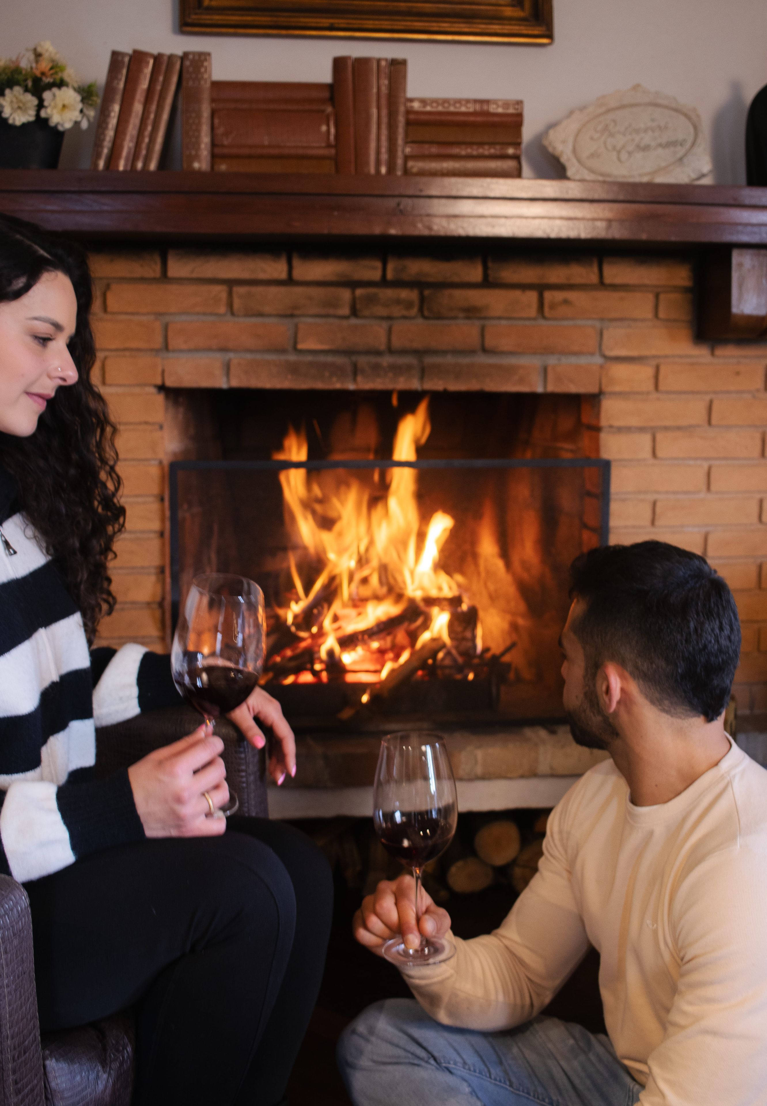 A couple seated in front of a fireplace with a fire burning, holding wine glasses.