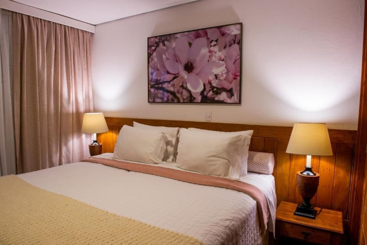 A hotel room with a bed, two bedside lamps, a large floral artwork on the wall, and a window with curtains.