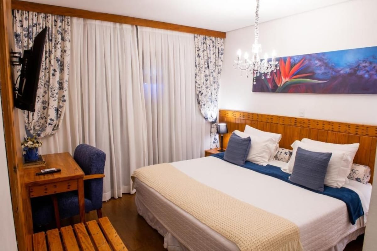 A hotel room with a bed, a desk with a chair, a television, and a painting on the wall.
