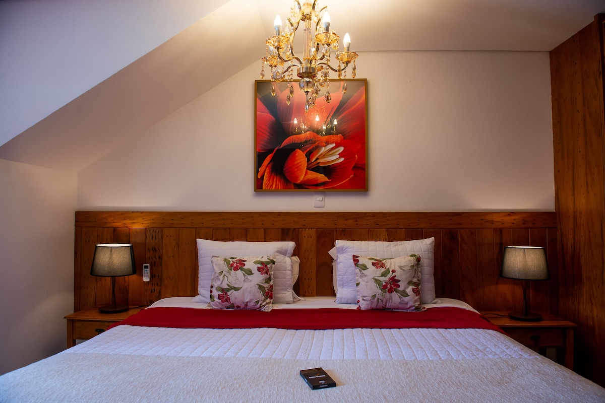 A bedroom with a wooden headboard, a chandelier, a painting of a flower, two bedside lamps, and a red throw blanket.