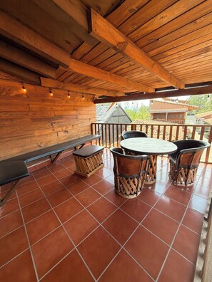 An outdoor patio area with wooden flooring, a round table with chairs, a bench, and string lights.