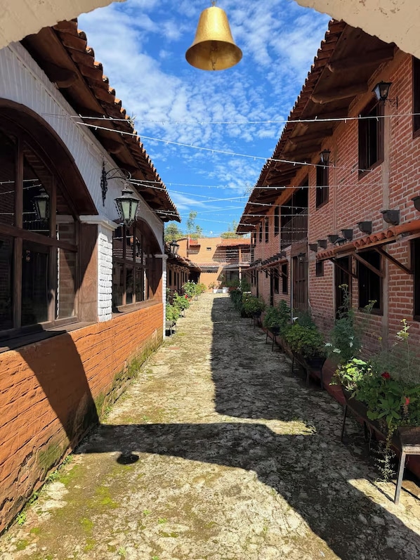 A narrow alleyway with brick buildings, a bell hanging overhead, and potted plants along the path.