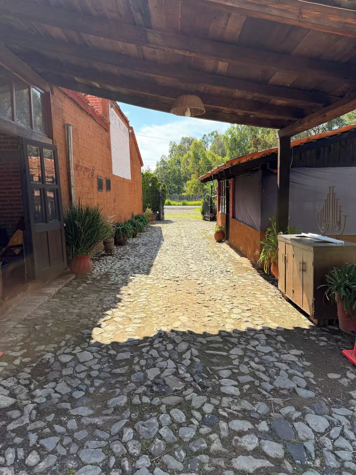 Un patio empedrado con edificios rústicos, una pérgola de madera y plantas en macetas.