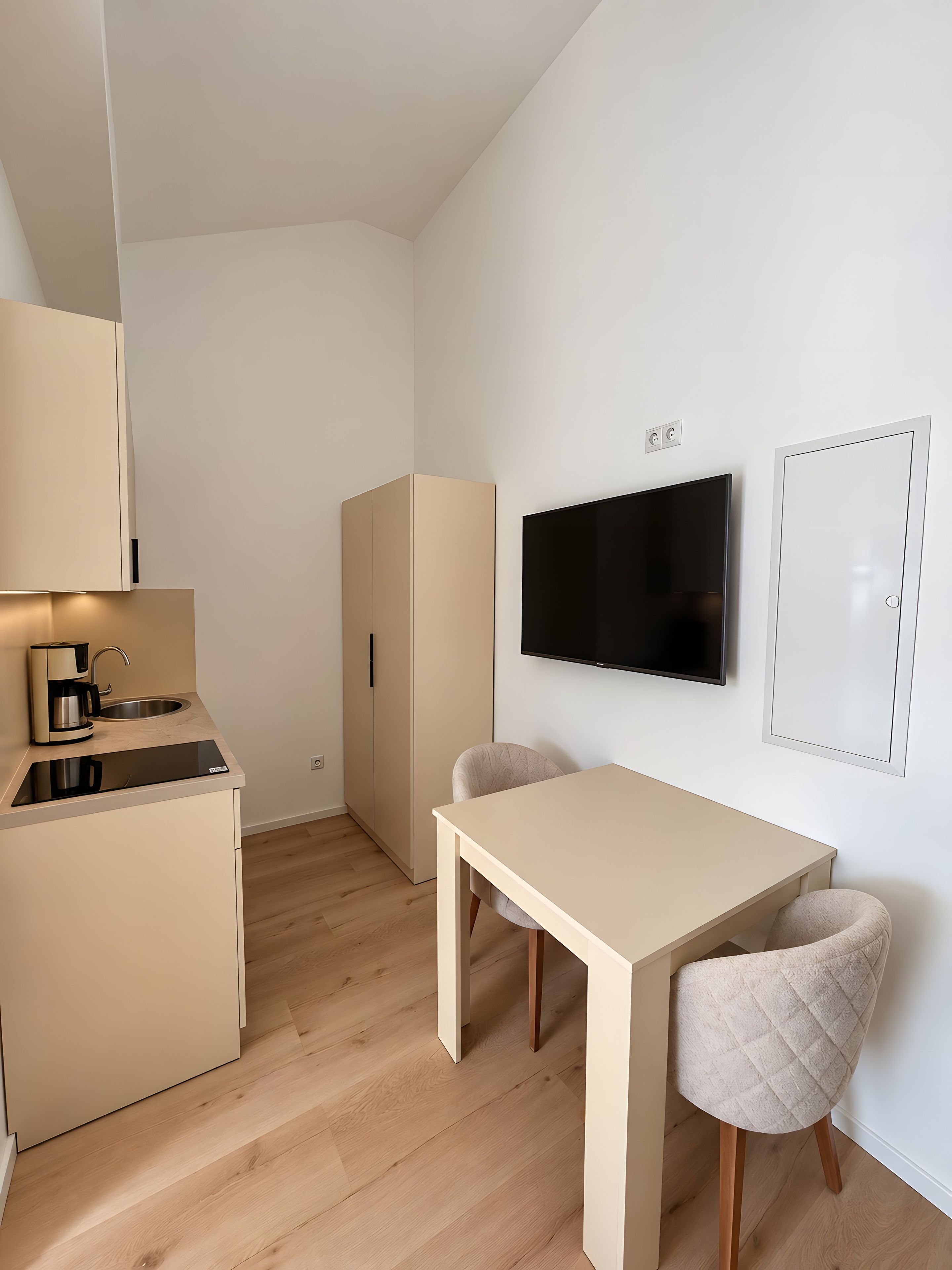A compact kitchenette with a coffee maker, a flat-screen TV mounted on the wall, and a table with two chairs.