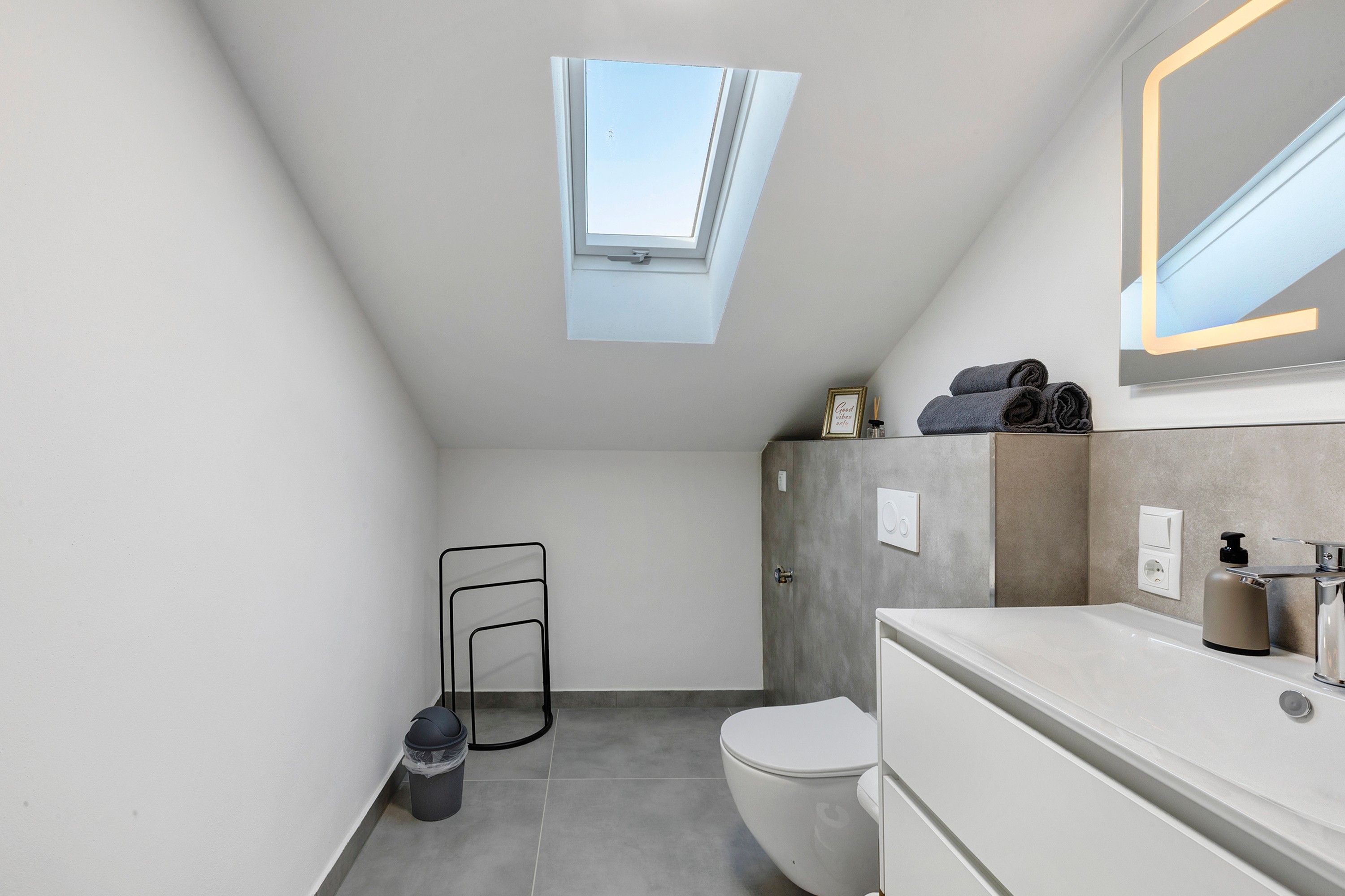Een moderne badkamer met dakraam, toilet, wastafel met spiegel en handdoekhouder.