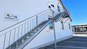 Un immeuble avec un escalier métallique menant à un balcon, une enseigne indiquant « APPARTEMENT 13 », et une zone de stationnement avec une enseigne « PARCOMÈTRE ».
