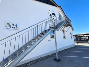 Un immeuble avec un escalier métallique menant à un balcon, une enseigne indiquant « APPARTEMENT 13 », et une zone de stationnement avec une enseigne « PARCOMÈTRE ».