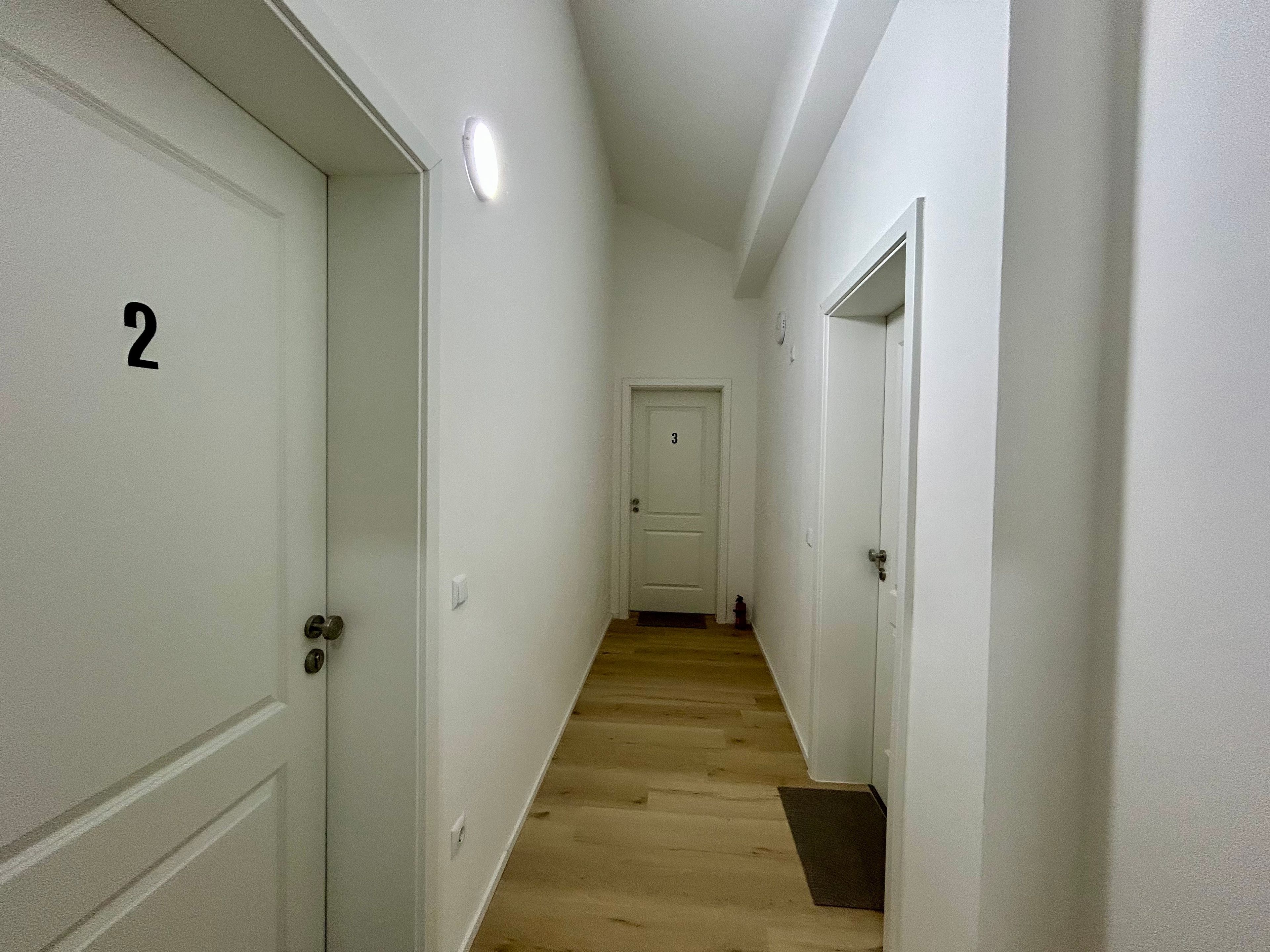 A narrow hallway with white doors numbered 2 and 3, wooden flooring, and a light fixture on the ceiling.