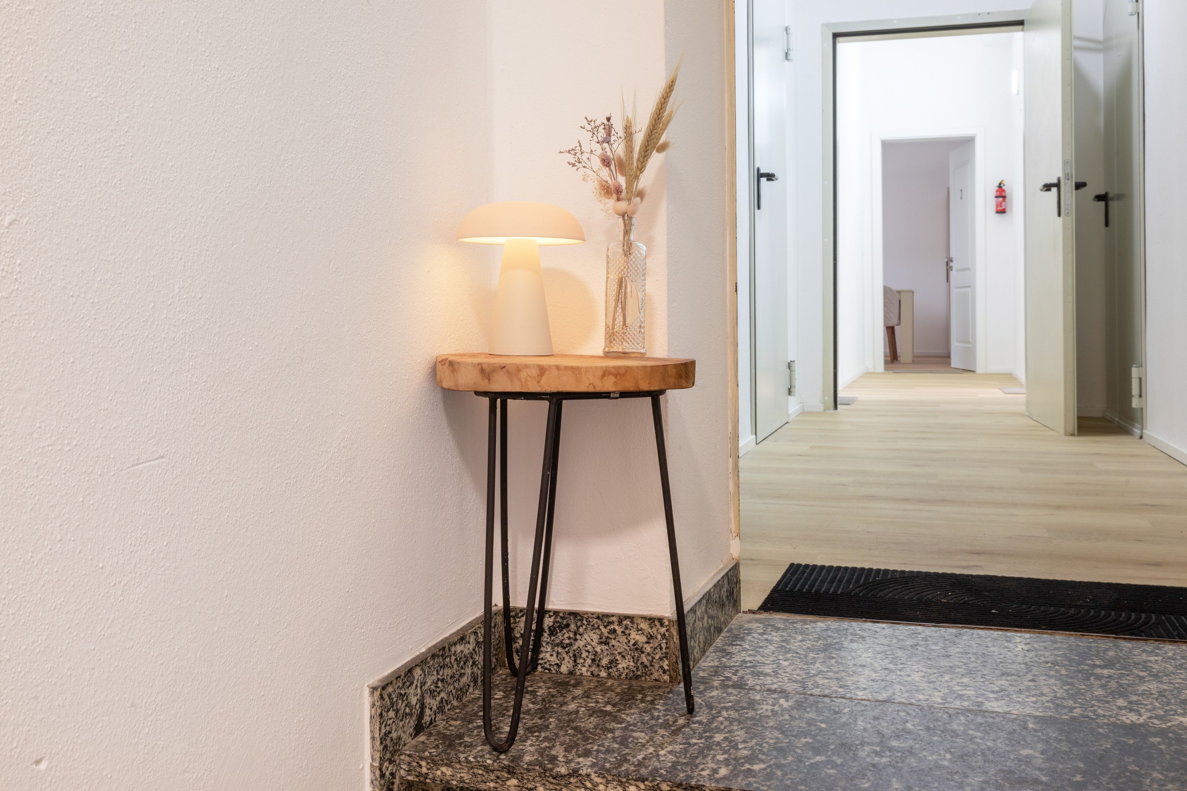 Een gang met een houten bijzettafeltje, een lamp en een vaas met gedroogde planten.