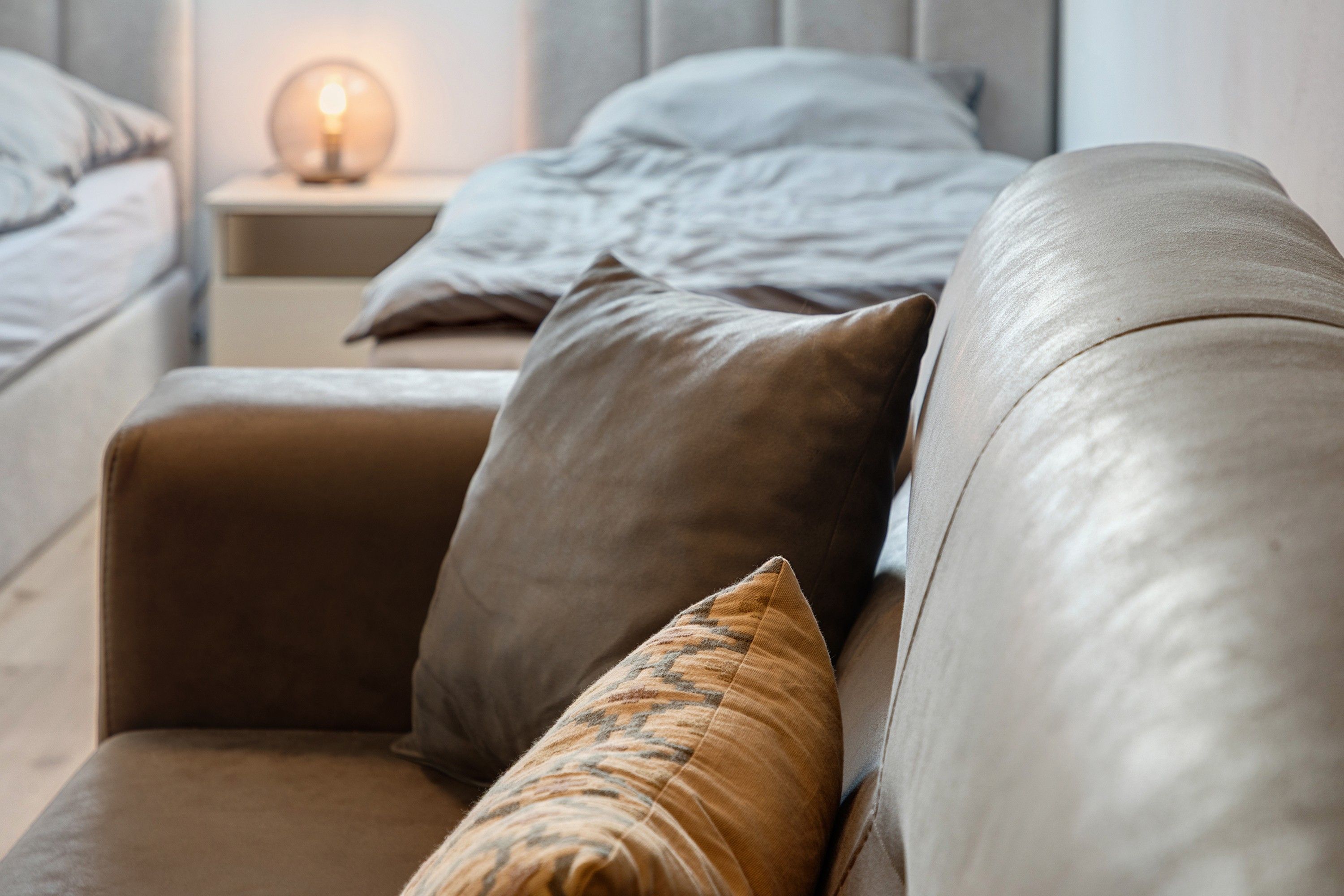 Ein modernes Schlafzimmer mit einer Ledersofa, einem Nachttisch mit Lampe und einem Bett mit grauer Bettdecke.