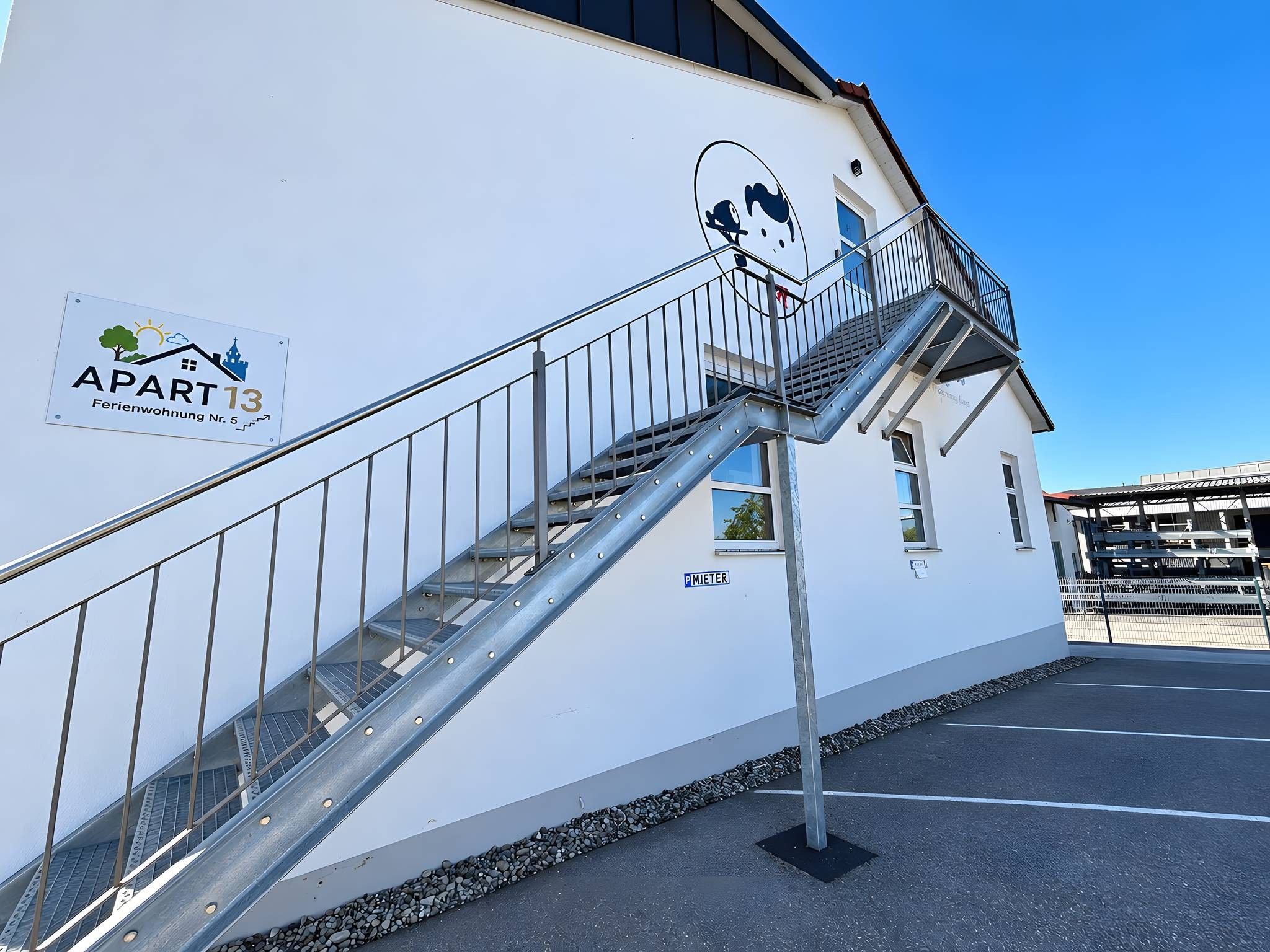 Un immeuble avec un escalier métallique menant à un balcon, une enseigne indiquant « APPARTEMENT 13 », et une zone de stationnement avec une enseigne « PARCOMÈTRE ».