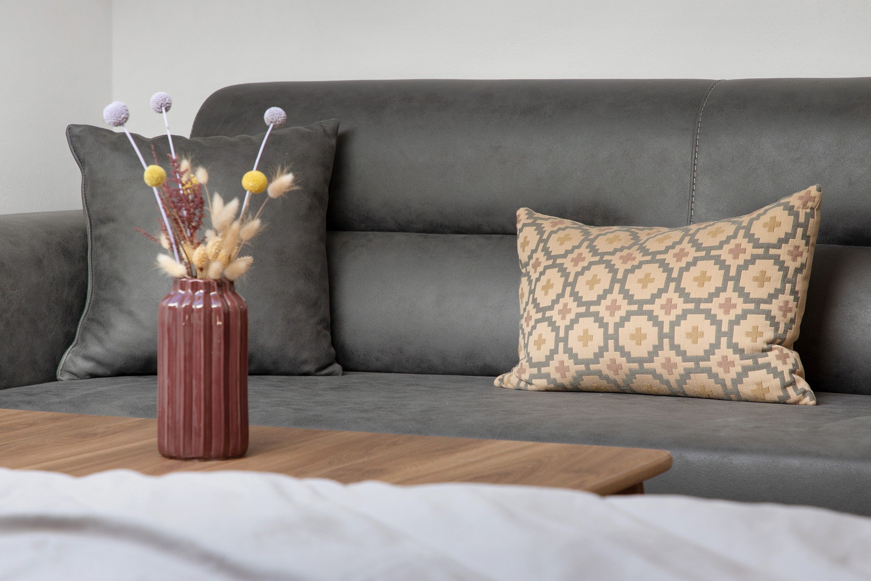 Ein graues Sofa mit einem gemusterten Kissen und einer Vase mit Trockenpflanzen auf einem Holztisch.