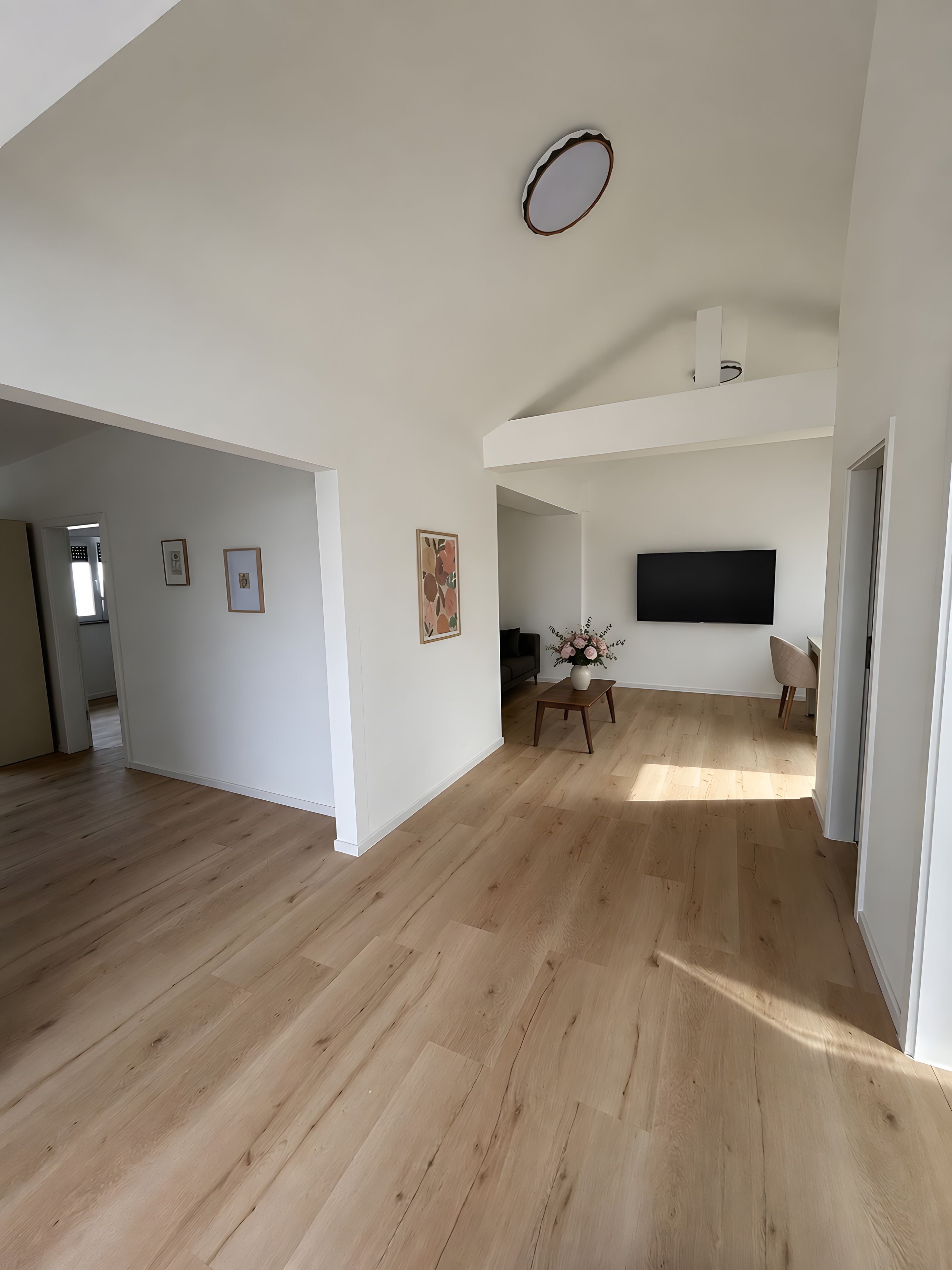 A spacious living area with wooden flooring, a flat-screen TV mounted on the wall, and a small table with a vase.