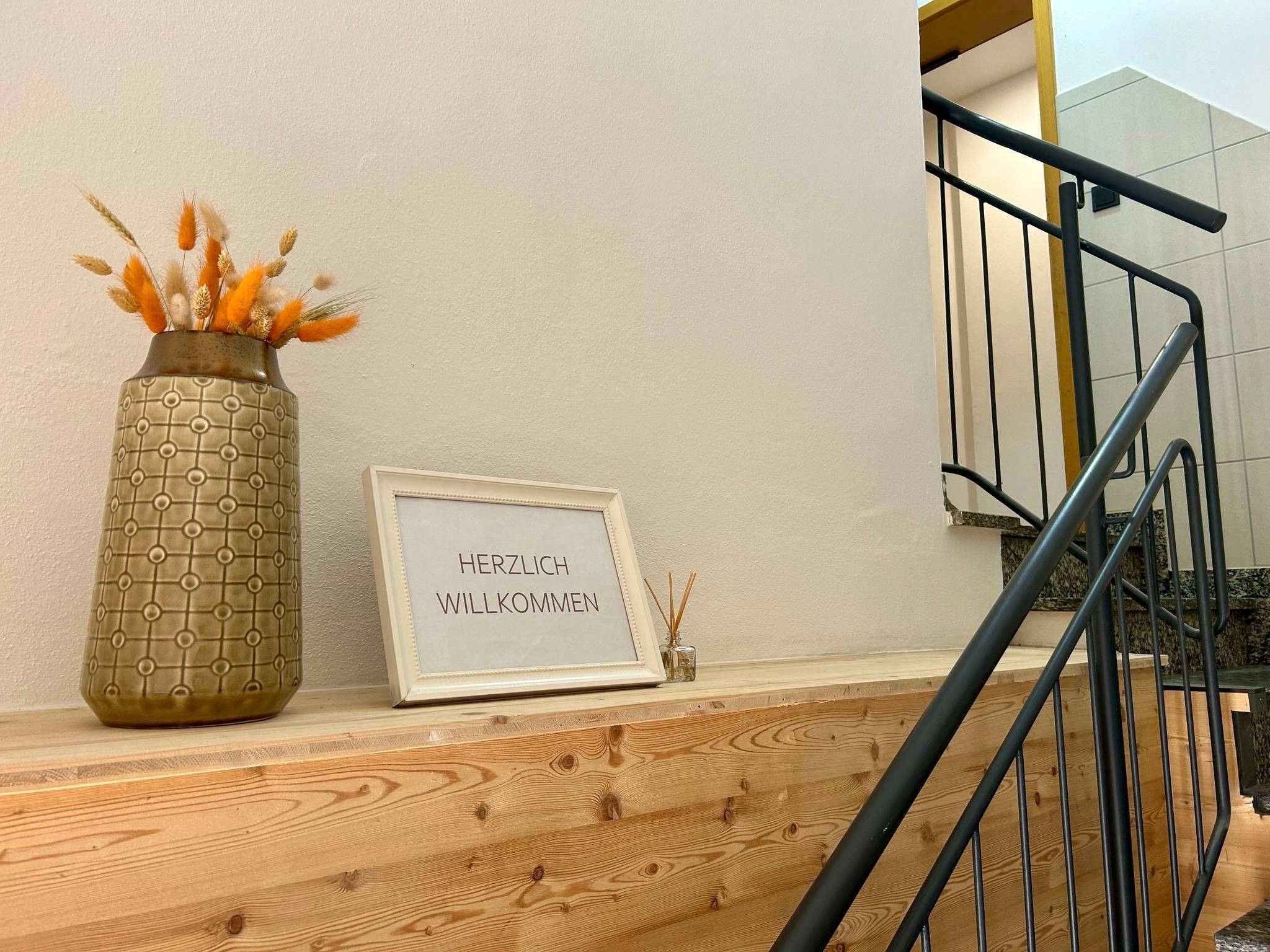 A wooden staircase with a metal railing, a vase with dried plants, and a framed sign with text.