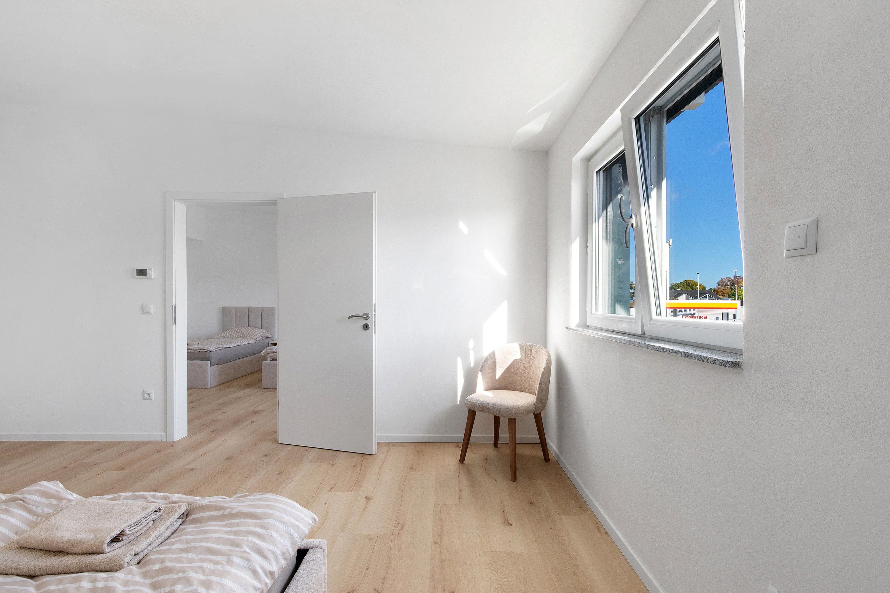 Ein modernes Schlafzimmer mit großem Fenster, einem Bett mit gestreifter Bettdecke, einem Sessel und Blick auf eine Tankstelle.