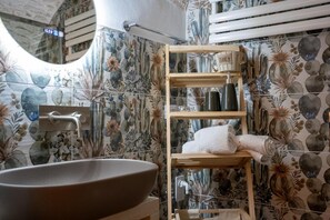 Un baño con una bañera independiente, un estante de madera con artículos de aseo y una pared con estampado floral.
