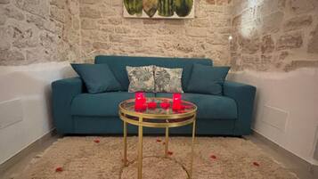 A room with a stone wall, a blue sofa, a glass coffee table with red objects, and a carpet with red petals.
