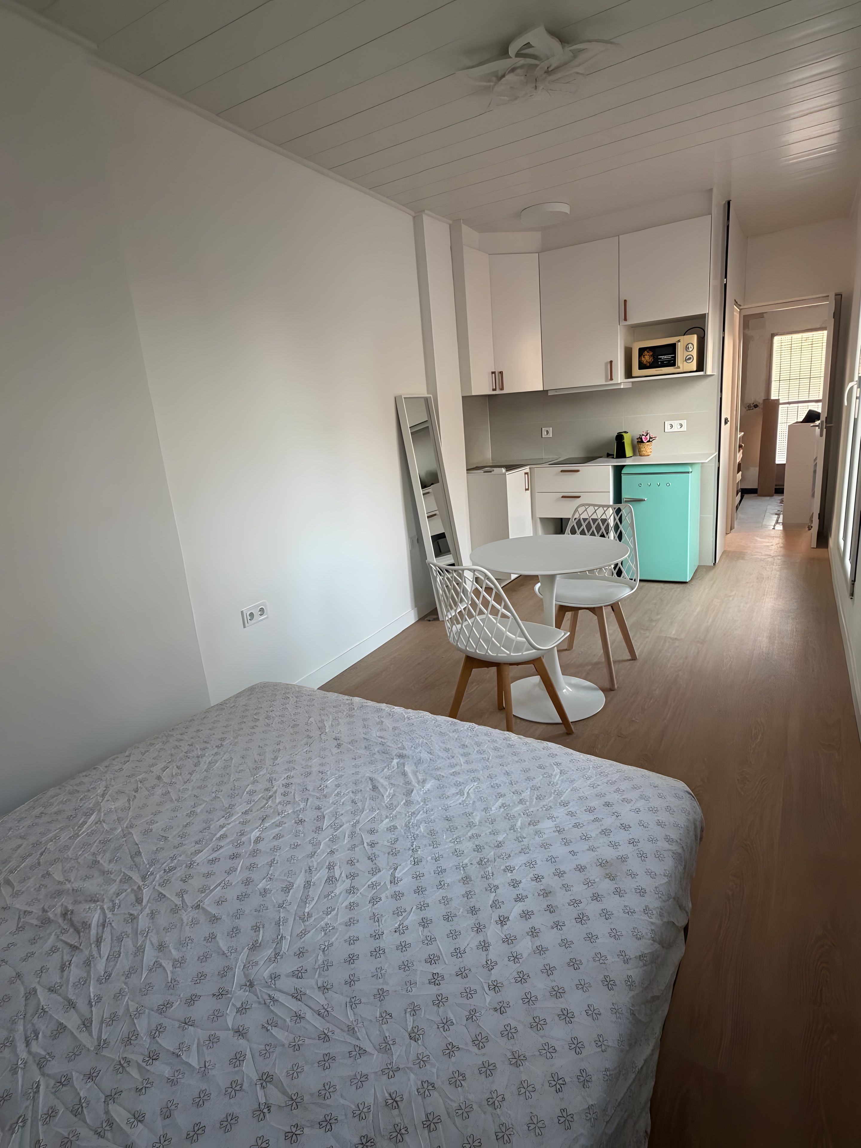 Een slaapkamer met een bed, een stoel, een bureau en een kitchenette op de achtergrond.