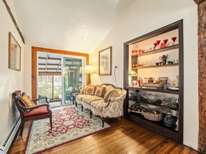 Un salón acogedor con piso de madera, un sillón rojo, un sofá estampado y un ventanal que da al exterior.