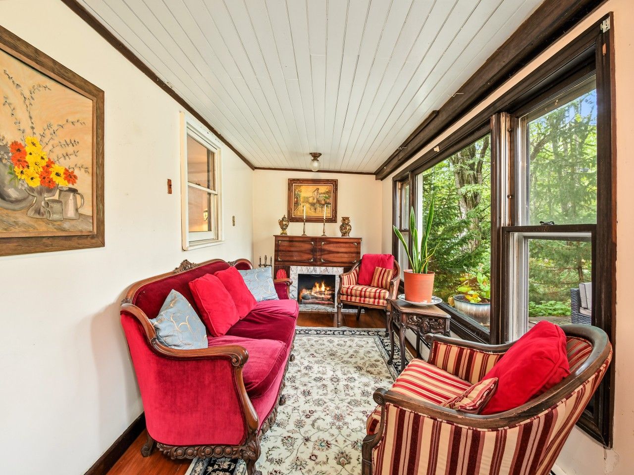 A cozy living room with a fireplace, red and striped furniture, and large windows offering a view of greenery.