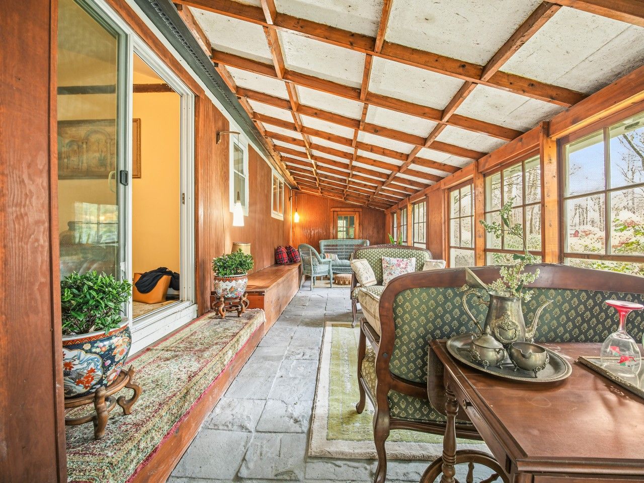 A covered porch with wooden beams, seating, potted plants, and a view of the outdoors.