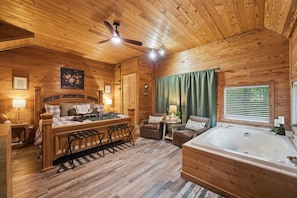 A wooden cabin interior with a bed, two chairs, a table, a ceiling fan, a window with curtains, and a bathtub. - Hot Tub-Fireplace-Private-Honeymoon-Hiking-Relax (Sevierville)