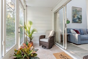 A sunlit room with a sliding glass door, a wicker chair, a blue sofa, and a potted plant. - Gator Glam (Gainesville)