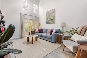 A modern living room with a sofa, coffee table, and armchair. There is a window with curtains, a painting on the wall, and a lamp on a side table. - Gator Glam (Gainesville)