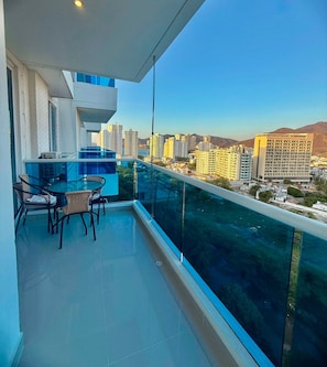 A balcony with a glass barrier, a dining table with chairs, and a view of a cityscape with buildings and mountains. - Reserva del Mar (Santa Marta)