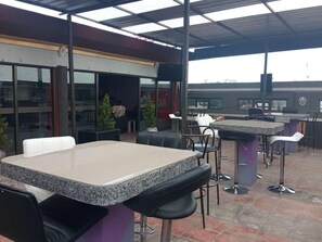 Un área de patio al aire libre con mesas y sillas, una sección cubierta con techo, y vista del edificio al fondo.