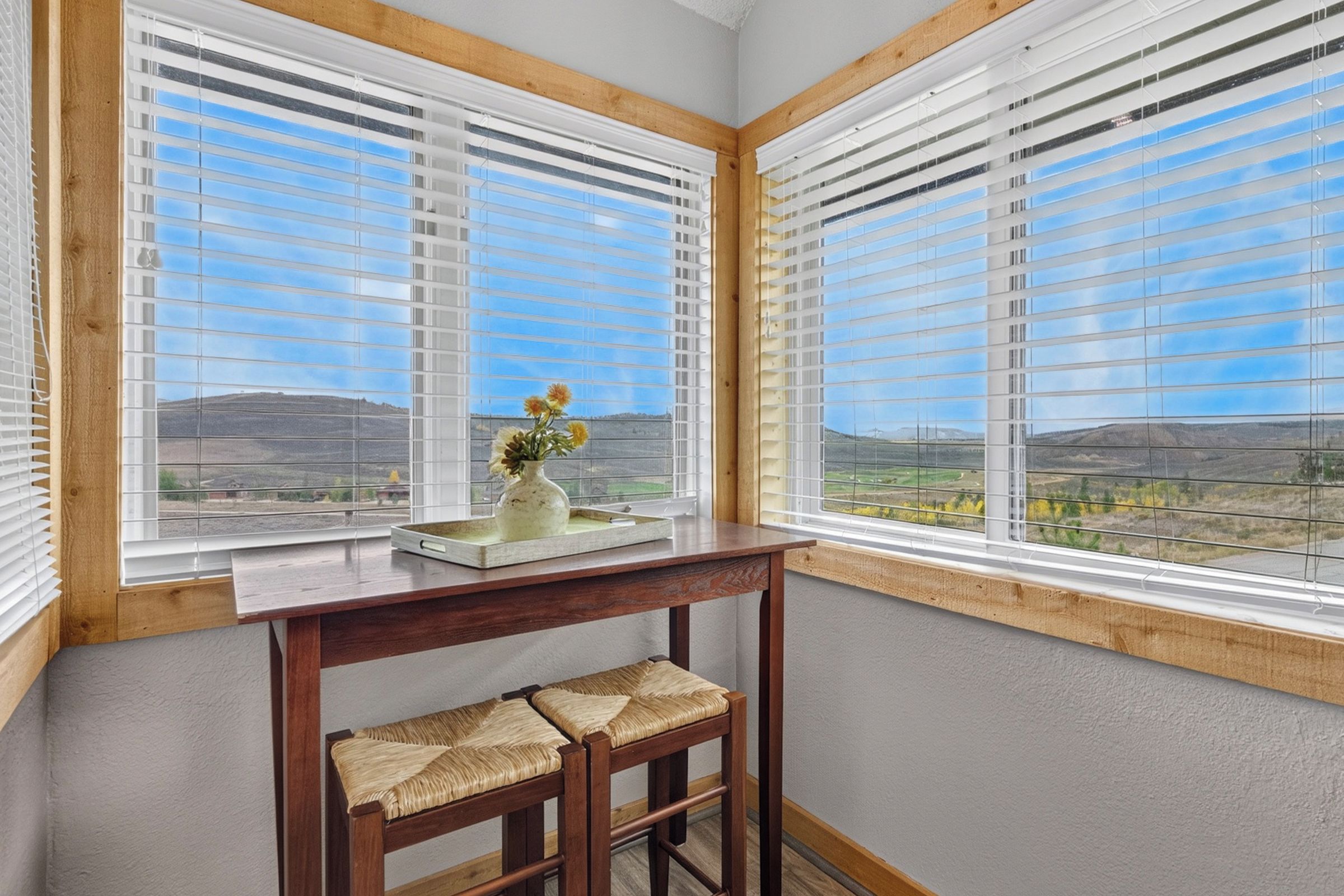 Una habitación de esquina con una mesa de madera y dos sillas, un jarrón con flores y vistas al paisaje a través de amplias ventanas con persianas.