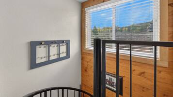 A room with a wooden floor, a window with blinds, a wall-mounted light switch panel, and a black metal railing.