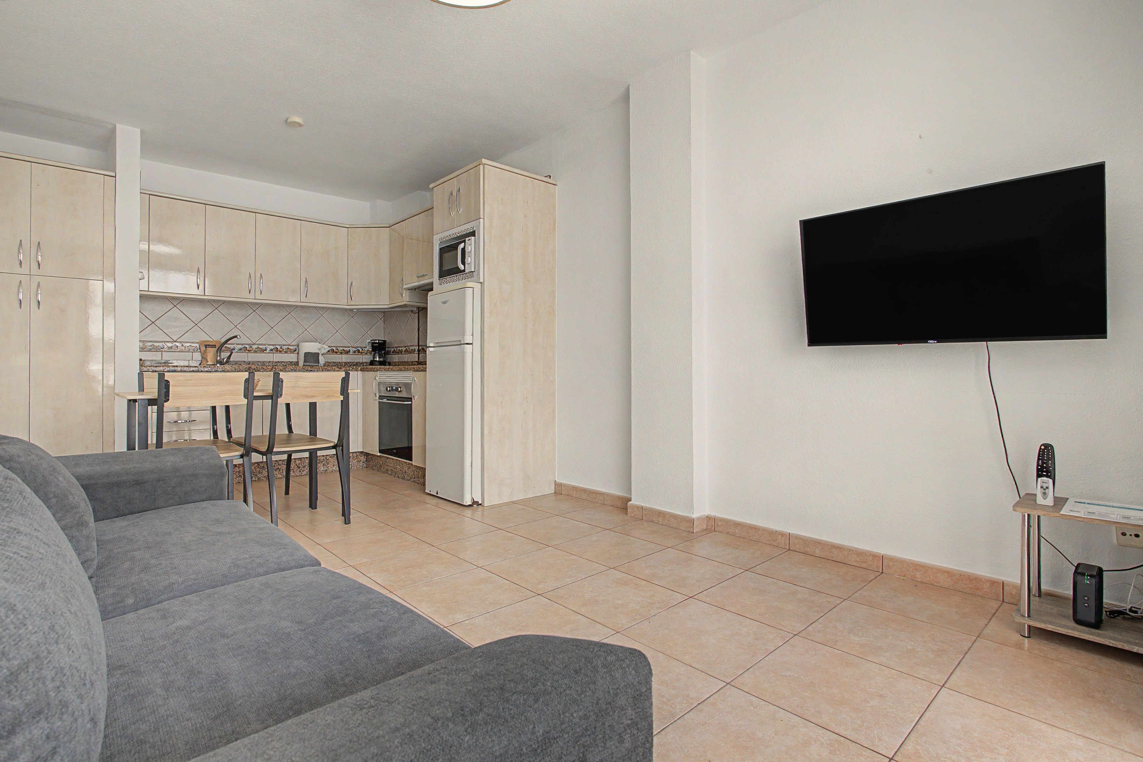 Een moderne woonkamer met een grijze bank, een flatscreen televisie aan de muur en een keuken met kasten en apparatuur.