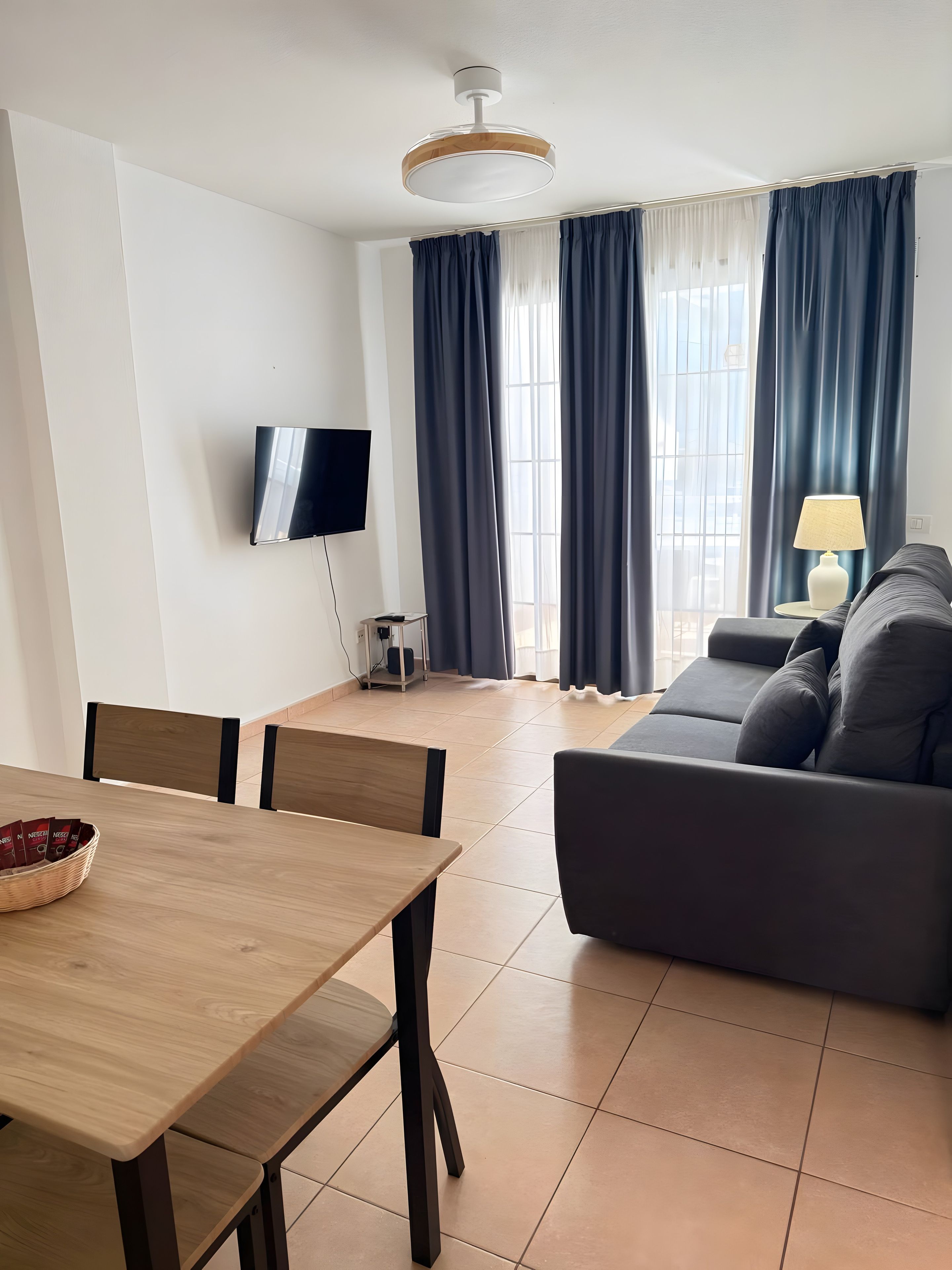 Un salon moderne avec un canapé, un téléviseur fixé au mur, une table à manger avec des chaises et une fenêtre avec des rideaux.