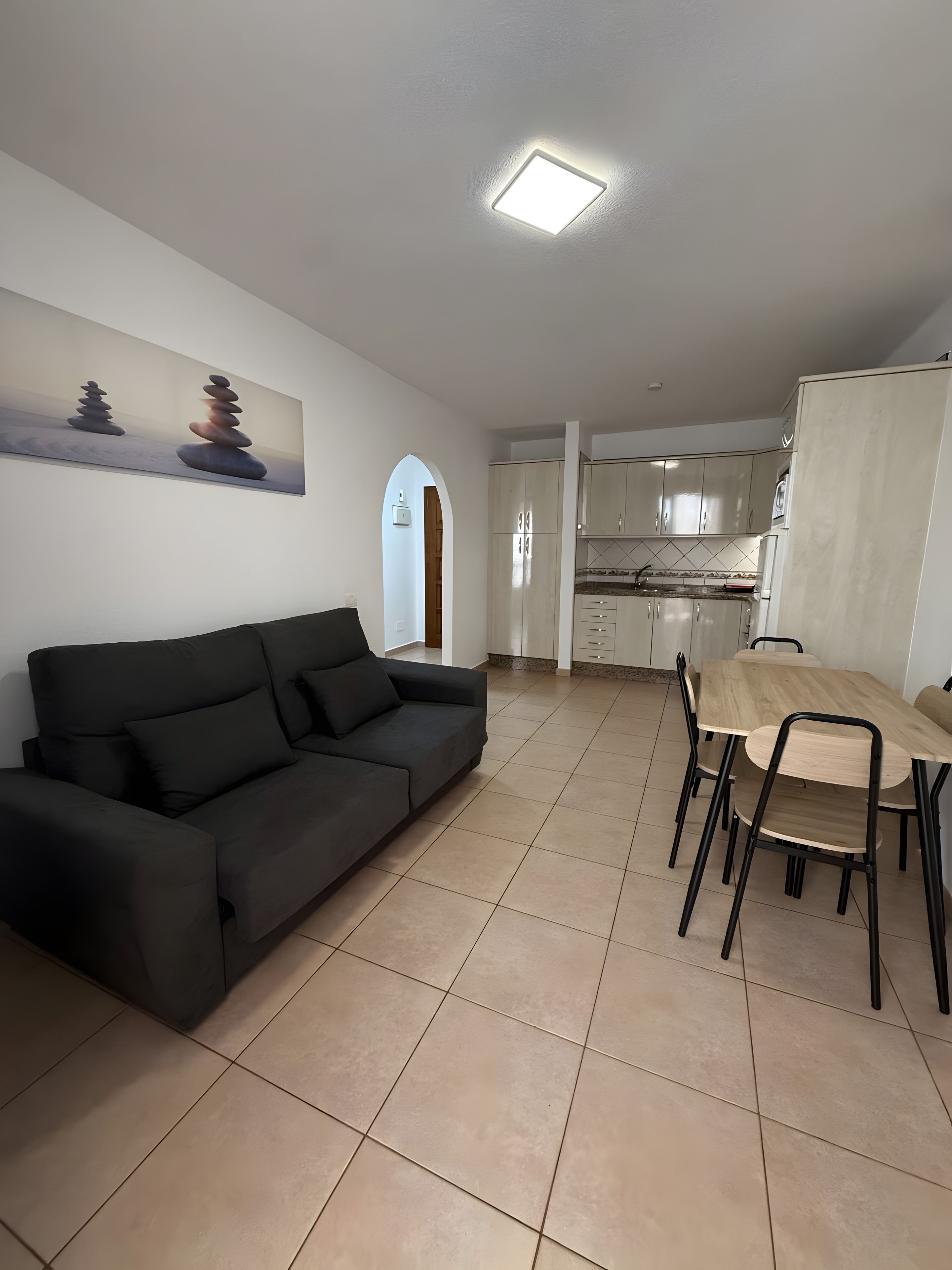 Un salon moderne avec un canapé noir, une table à manger avec des chaises et un coin cuisine équipé de placards et d’un réfrigérateur.